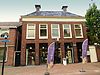 Vijf traveeën breed pand met verdieping en forse kroonlijst onder schilddak met twee hoekschoorstenen waarop borden