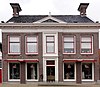 Groot pand met omlijste ingangspartij met snijwerk en timpaan met piron, verdieping onder dwarskap tussen zijtopgevels en daarboven grote schoorsteen met dubbel bord en bekroning