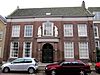 Nieuwstraat 12 - Oude Mannen- en Vrouwenhuis. Breed pand met verdieping en zadeldak. Breedte vijf vensterassen. De eenvoudige, bakstenen lijstgevel heeft in baksteen gebosseerde hoeklisenen en een risalerende middenpartij. Hierin de getoogde ingang, geflankeerd door bakstenen Dorische pilasters, die een hoofdgestel dragen. Te weerszijden van het door Lodewijk XVI-snijwerk met siervaas bekroonde venster boven de ingang de stenen beelden van een oude vrouw en een oude man. Hardstenen plint; vensters met 15- en 9-…