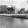 Overzicht voor- en zijgevel boerderij met leilinden voor het woonhuis, waterpartij op voorgrond