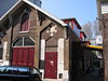 WLM - Minke Wagenaar - 126 2660 - Omschrijving rijksmonument Bakstenen remisedeel met de voorgevel aan de Schinkelhavenstraat 27 en de achtergevel aan de Eerste Schinkelhavenstraat 16. Aan de oostzijde grenst het gebouw aan het binnenterrein. De voorgevel geeft de onderverdeling van dit deel van het gebouw in drie bouwlagen weer: hoofdverdieping, verdieping en kapverdieping. Op de hoofdverdieping een brede toegangsdeur met luifel. Onder de overkapping van de luifel de initialen van de A.O.M. tussen florale decoratie. Ter rechte…