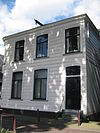 Gedeeltelijk houten en gedeeltelijk bakstenen dijkhuis met lijstgevel in empirestijl uit de 19de eeuw, overhoeks geplaatst gebogen ramen