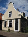 Houten dijkhuis met klokgevel (uit- en ingezwenkt voorschot) uit 18de eeuw. Onderpui in empirestijl, puntgevel aan achterzijde
