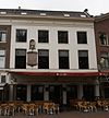 Geschilderd pand met brede gevel met rechte kroonlijst met tandingen. Beneden verbouwd