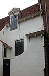 Witgepleisterd laag huis met hoog zadeldak en uitbouwen. Dakkapel door daklijst snijdend met hijsbalk