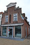 Hoekpand met afgeknotte trapgevel met klauwstukken. Gecementeerde gevel met schuiframen. Stoep met mooie zijhekken