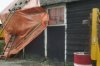 Voorgevel van de boerderij, detail: deur en de gedeeltelijk afgebroken zijgevel, met oranje bouwzeil