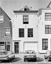 Huis met gepleisterde lijstgevel, aan de hoeken ingezwenkt. Dakkapel. Voordeuromlijsting. Aan de achterzijde een ontpleisterde trapgevel