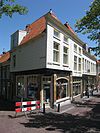 Gepleisterd huis onder schilddak. dat achter aansluit tegen een puntgevel. Aan de voorzijde een gepleisterde lijstgevel met lage bovenverdieping